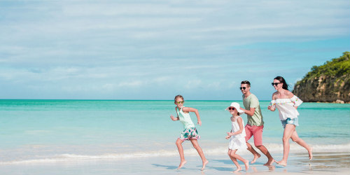 Boldog család, azaz fiatal apa, anya, egy kisfiú és egy kislányt vidáman fut a tengerparton, mezítláb az éppen visszahúzódó tengervizes homokon, a háttérben egy sziget kis része látszik a türkiz vízben. 